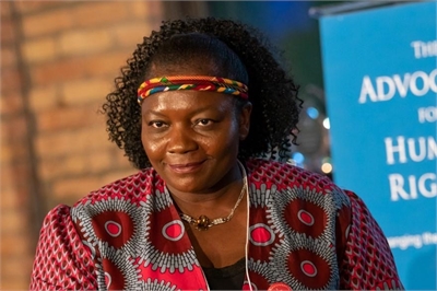 [ai] A woman with curly hair wearing a colorful patterned blouse and a decorative headband, looking directly at the camera with a neutral expression, in front of a blue banner with text related to human rights advocacy.